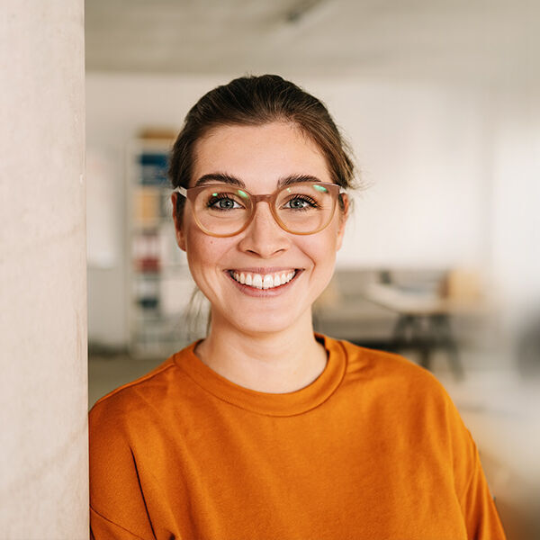 eine Frau in einem orangefarbenen Hemd und einer Brille, die an eine Wand gelehnt ist